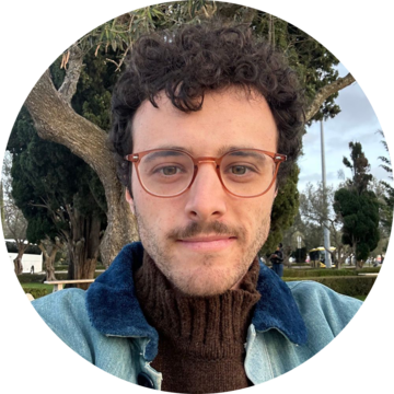 Adam Mahler Portrait - Curly haired person with mustache, red glasses, and blue denim jacket with blue sherpa cotton collar 